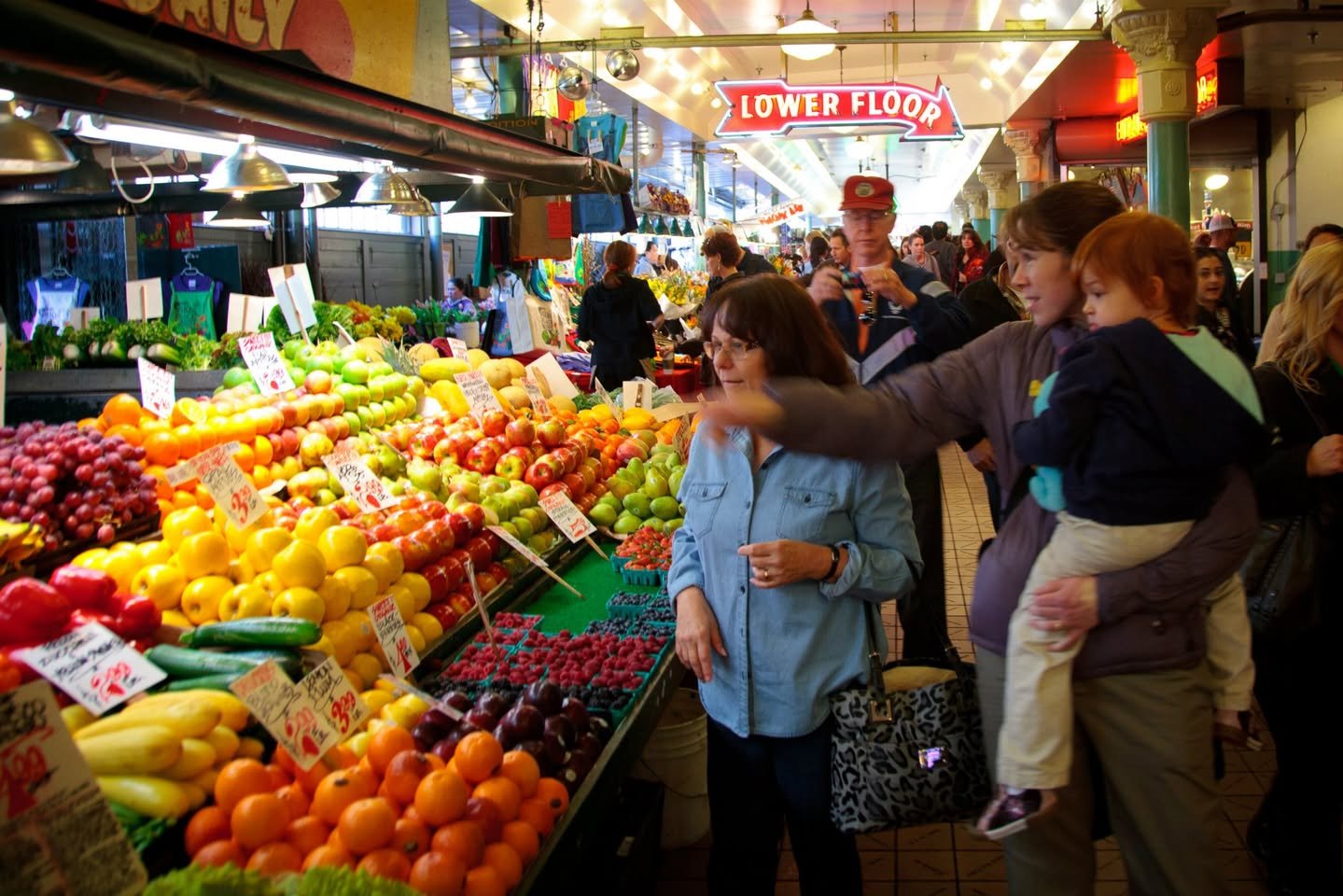 A group of people browsing a colorful fruit stand in a bustling indoor market under a 'Lower Floor' sign.