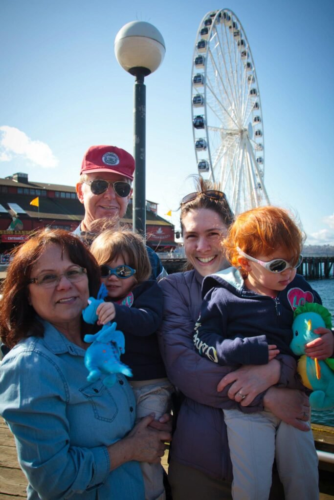 A family poses for a photo in front of the Seattle Great Wheel at the waterfront. The scene includes a large Ferris wheel under a clear blue sky.