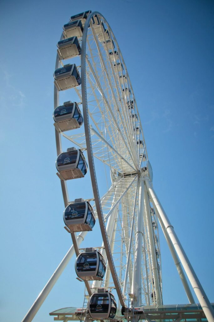 A tall Ferris wheel with enclosed passenger cabins stands against a bright blue sky, captured in multiple angles.