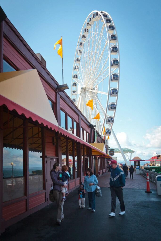 A large iron wheel with passenger cabins sits beside a red market building on a sunny day at Seattle's Pike Place Market.
