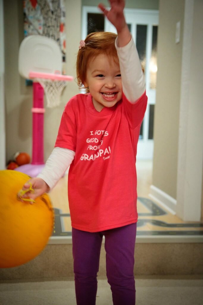 A young child in a red shirt smiles while holding an orange ball and raising one arm in a home setting.