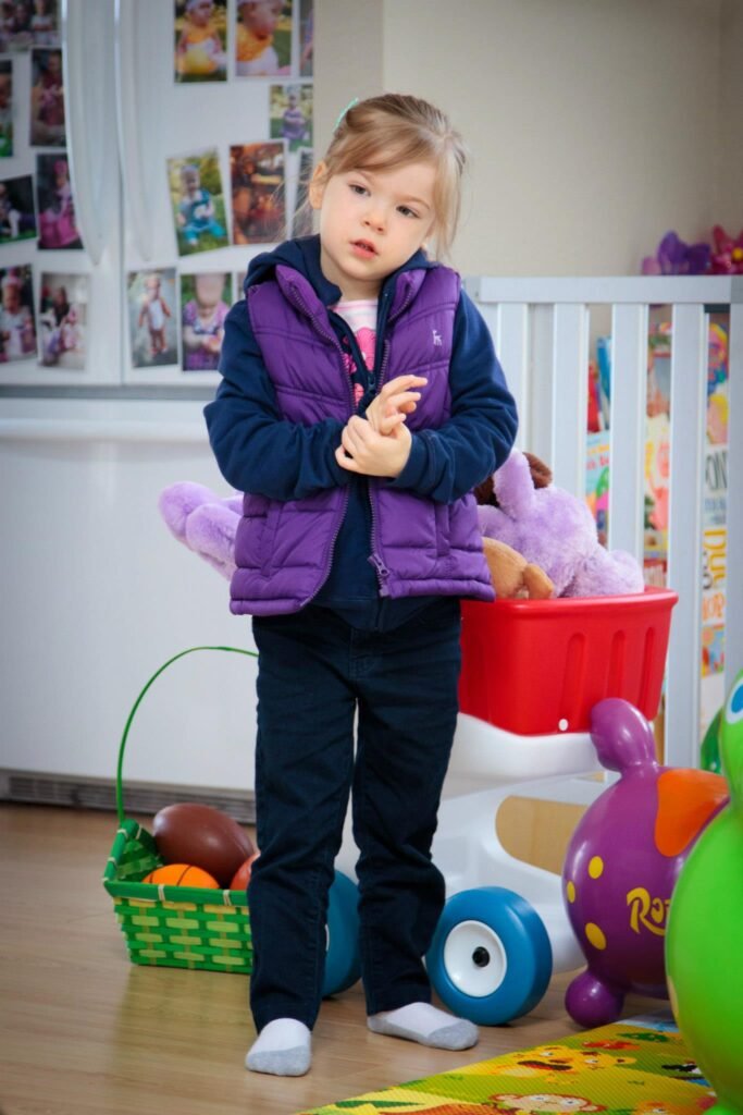 A toddler wearing a purple vest stands among toys in a bright playroom. Stuffed animals and colorful ride-ons surround the child on a wooden floor.