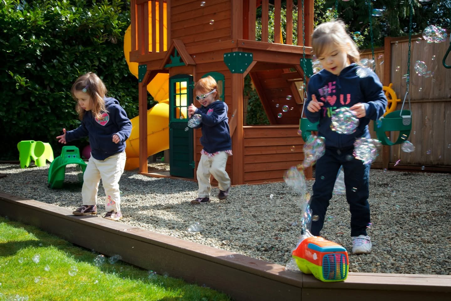 Children playing with bubbles in a backyard featuring a wooden playset.