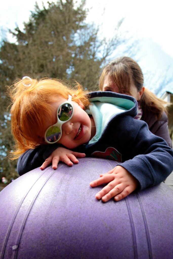 A smiling toddler in sunglasses plays on a purple exercise ball outdoors.