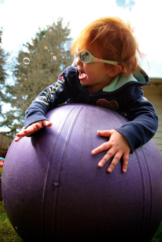 A young toddler with glasses is actively climbing on a large purple exercise ball outdoors.