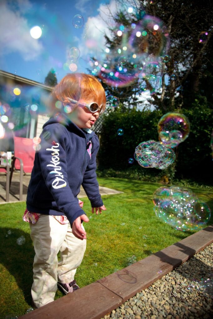 A young person in a blue hoodie plays with soapy bubbles in a sunny backyard with green lawn and gravel path.