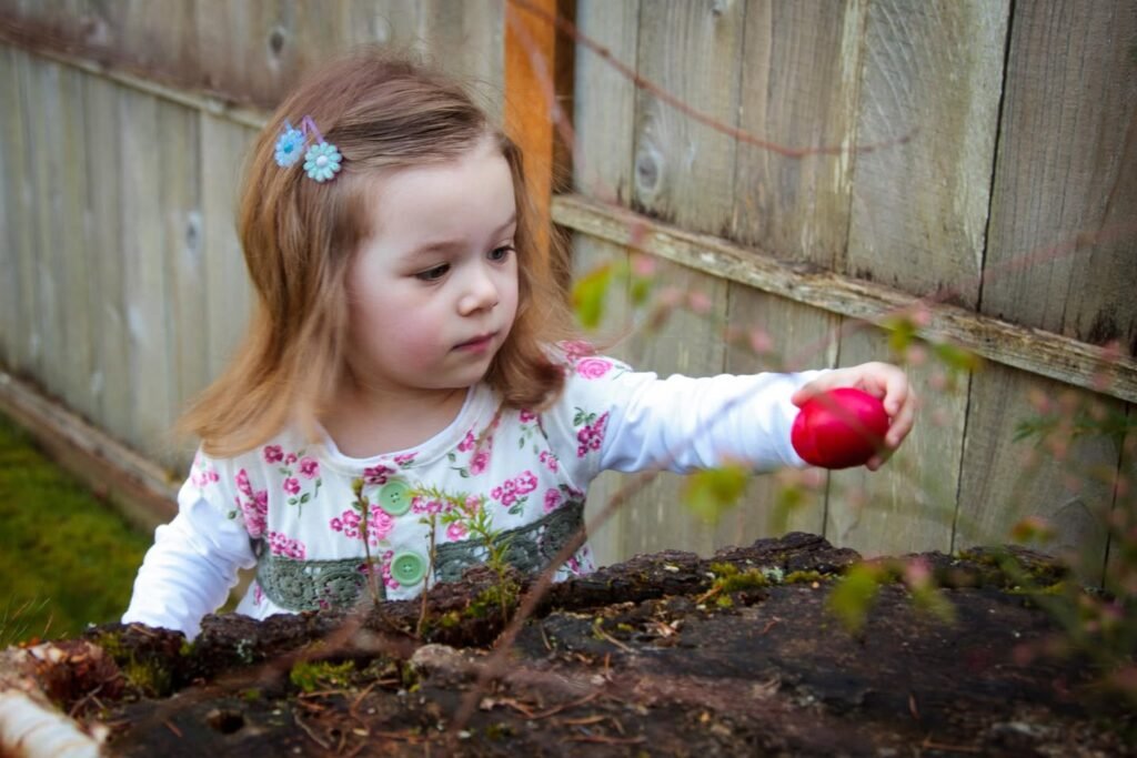 A young girl wearing floral clothes is placing a red object on the garden soil. She is standing in front of a wooden fence.
