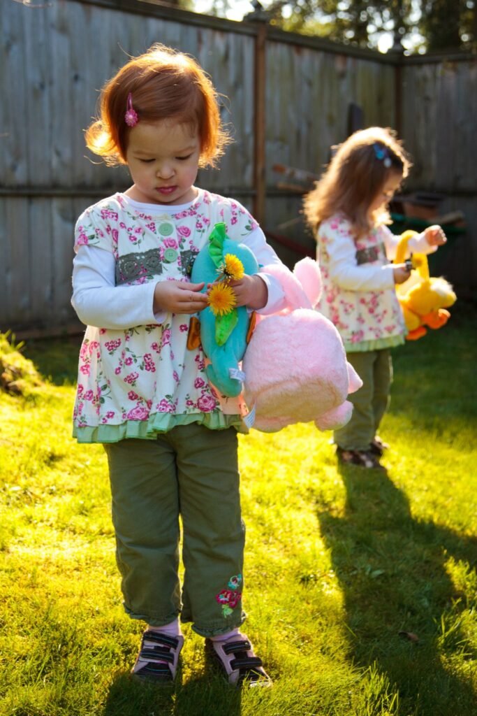 A fair-haired toddler in a floral dress plays with a pink stuffed animal on the grass.