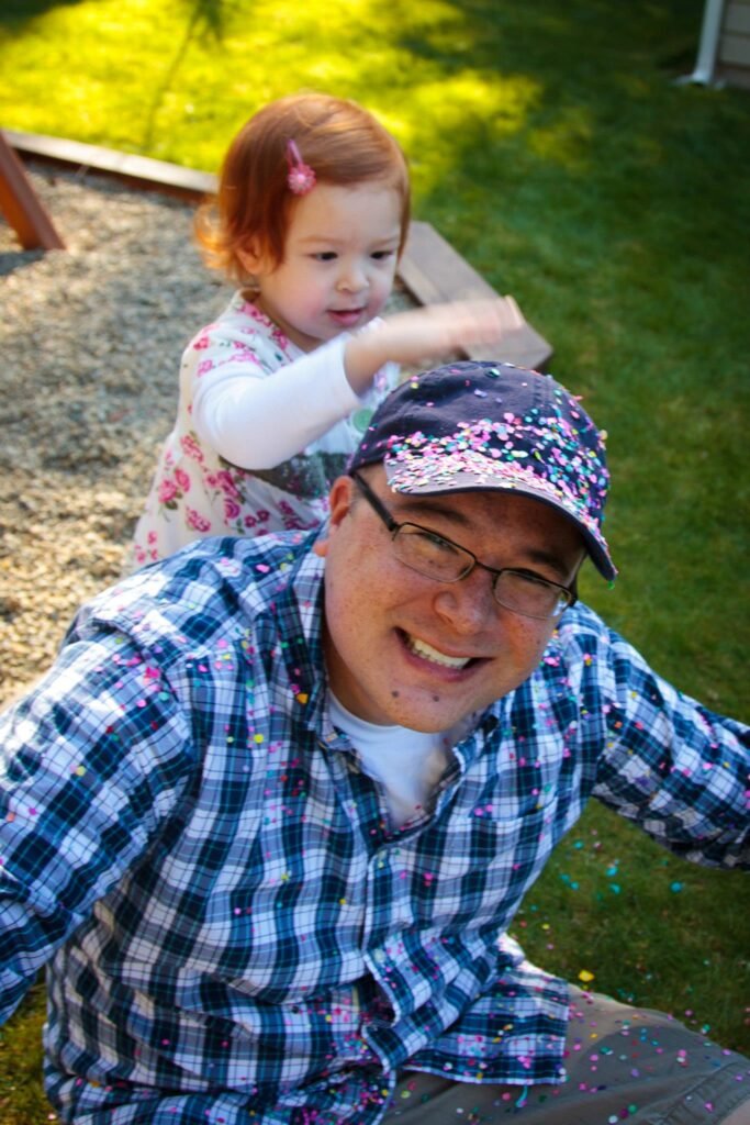 A smiling person in a cap and plaid shirt sits outdoors while a toddler playfully pats their head. The scene captures a joyful moment in a sunny backyard.