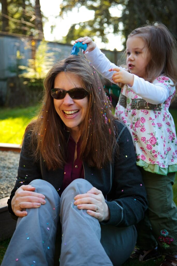 A smiling woman sits in a garden while a young child playfully sprays glitter on her hair.