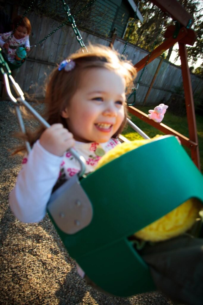 A smiling child enjoying a ride on a green bucket swing in a sunny backyard. The image captures a happy moment during outdoor play on a wooden swing set.
