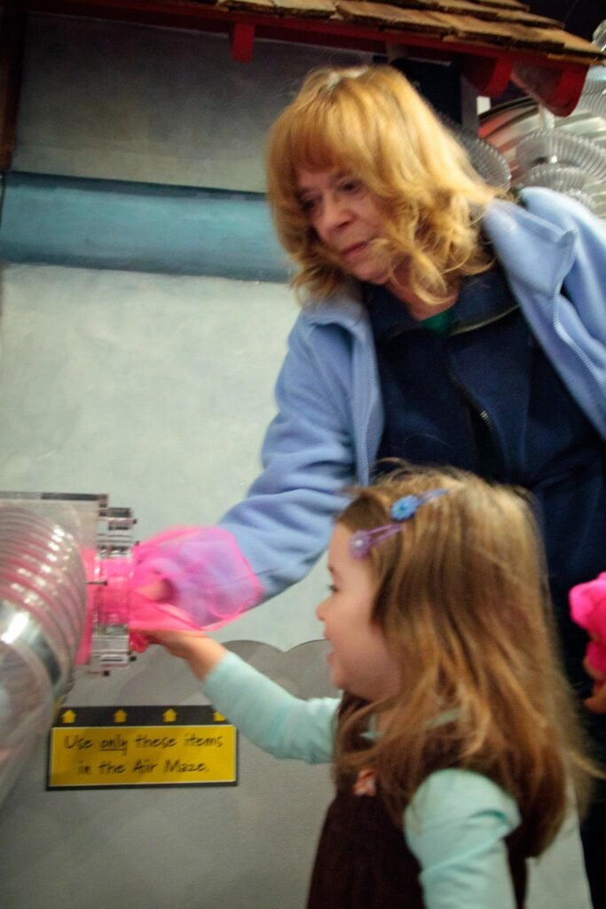 A person assists a child with a hands-on exhibit at an air museum. The child reaches towards a control panel labeled 'Use Only Small Feet'.
