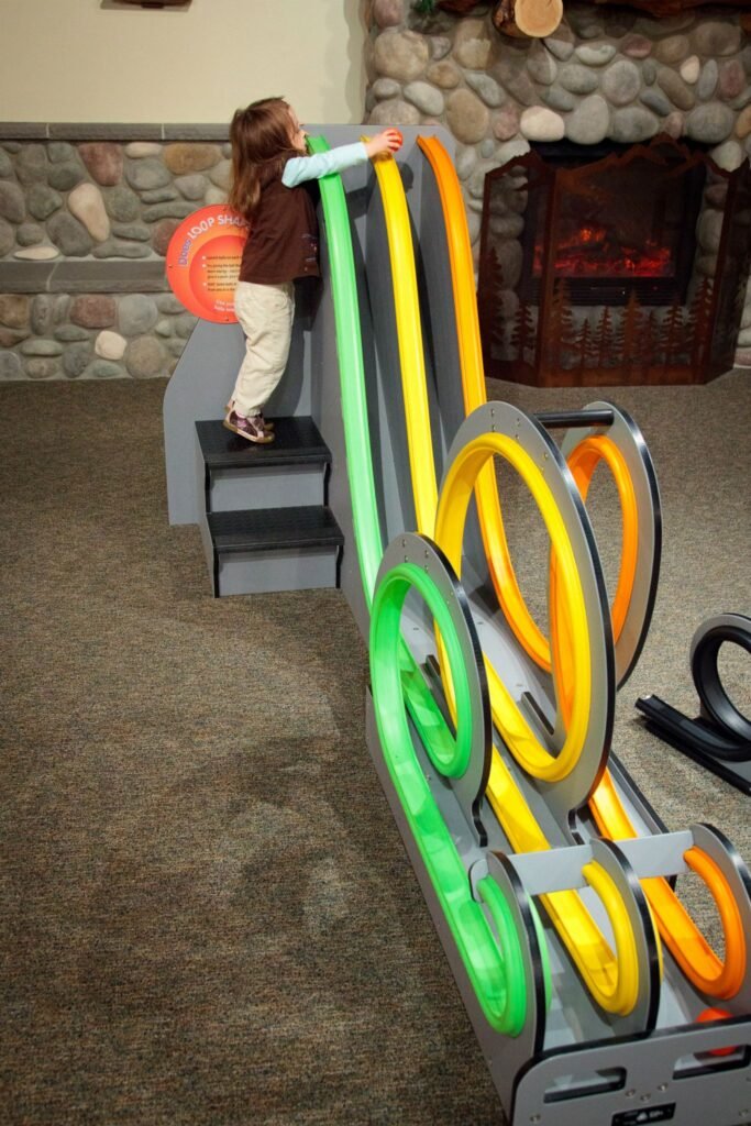 A customized indoor wooden marble run with colorful loops and stair access, located in a living room near a stone fireplace and rug.