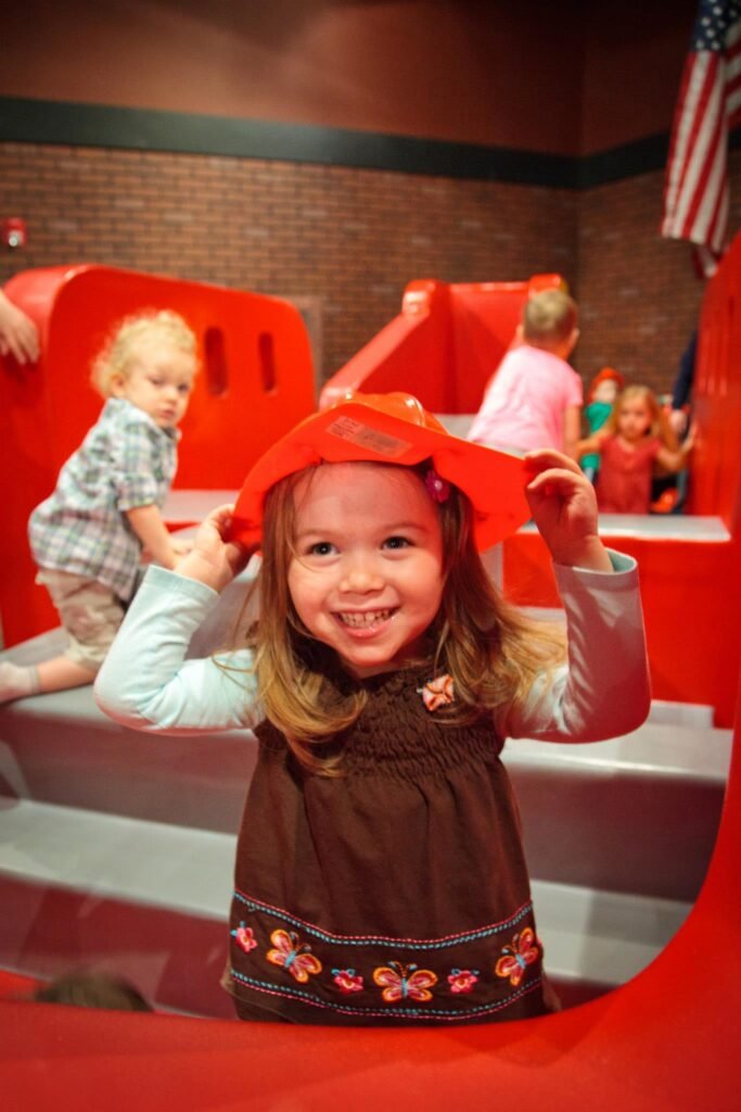 A child enjoys playing on a red fire truck exhibit in a museum with an American flag nearby.