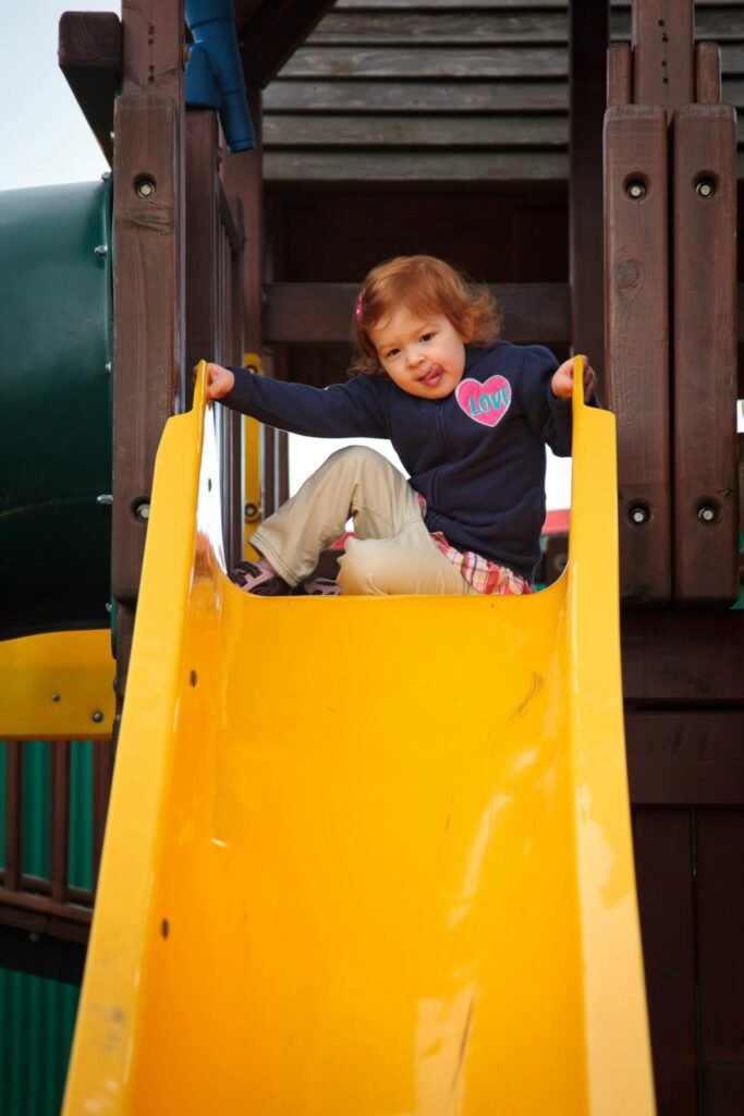 A toddler sitting at the top of a yellow playground slide on a wooden play structure.