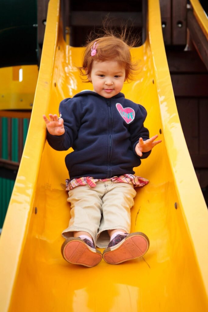A toddler in a navy sweater and patterned waistband sits on a yellow playground slide with an open mouth expression.