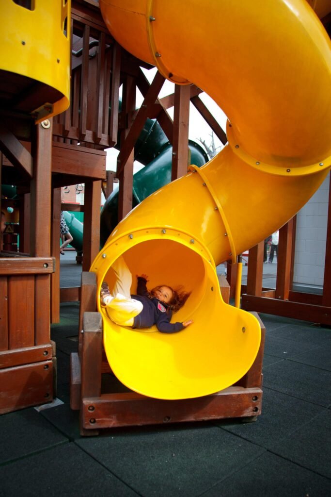 A bright yellow tube slide at an indoor playground. The structure is built with wooden beams and features playful design.