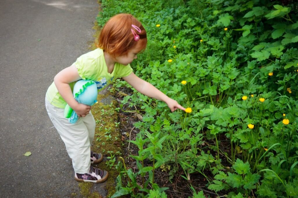 A red-haired child in a green shirt points to yellow flowers growing beside a paved path.