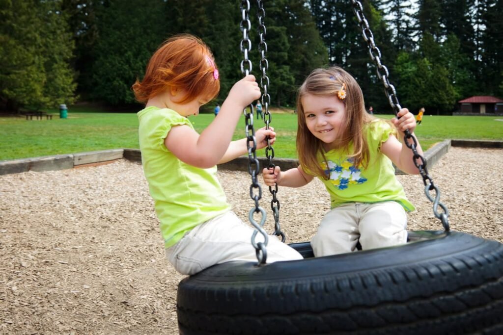 Two children enjoy playing on a tire swing at a local playground. The background features lush green trees and wood chip ground cover typical of a park setting.