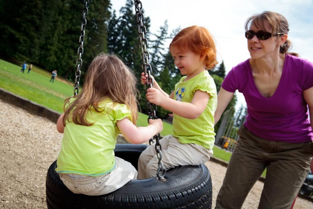 Two children swinging on a tire swing while a woman smiles on the side at a sunny outdoor park.
