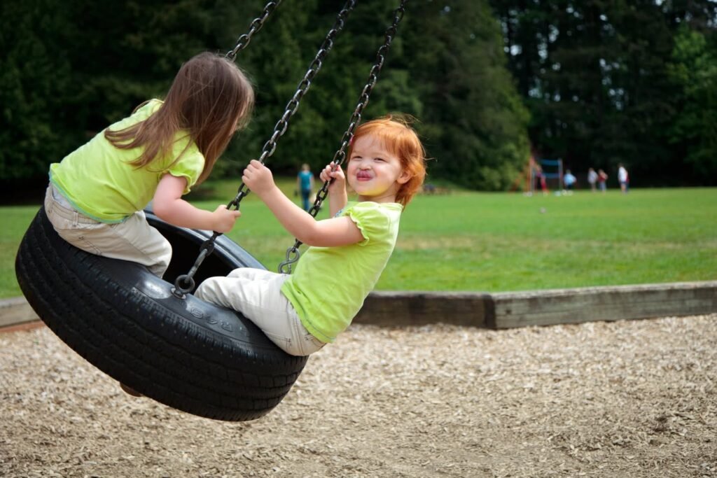 Two children in bright green shirts enjoying a tire swing in a sunny park with grass and trees.