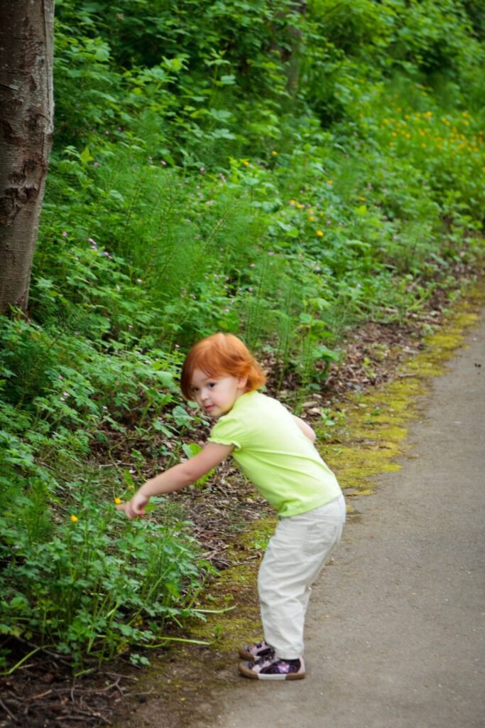 A child in a bright green shirt reaches for plants beside a paved path in a lush green forest setting, with views of a tree trunk and dense vegetation.