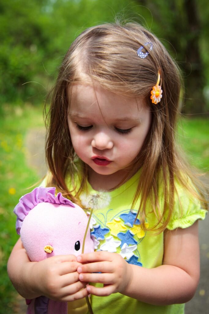 A young child in a yellow shirt gently inspects a wildflower while holding a pink plush toy. Bright hair accessories accentuate the cheerful Pacific Northwest setting.