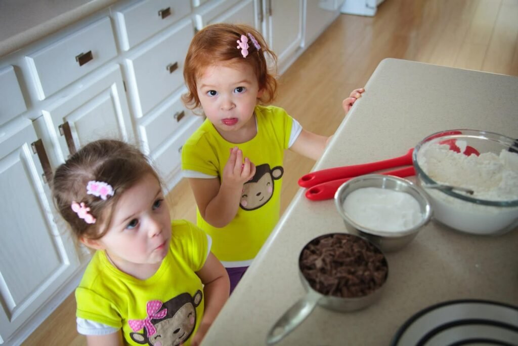 Two young children stand at a kitchen counter with bowls of flour and chocolate chips, wearing matching yellow shirts.