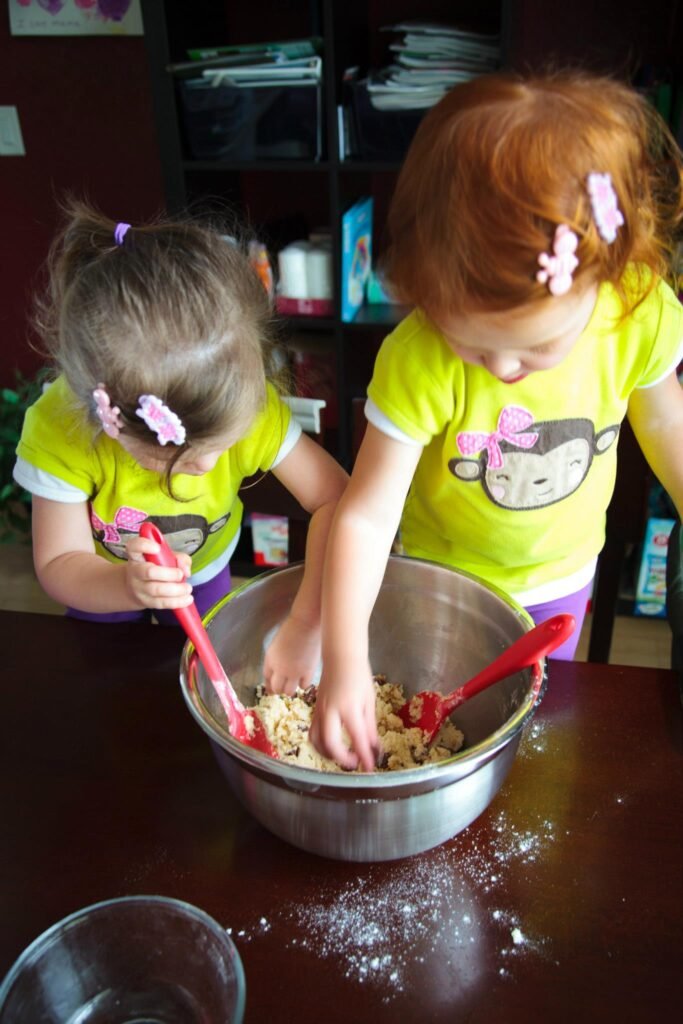 Two children in matching yellow shirts are mixing ingredients in a large metal bowl on a kitchen table.