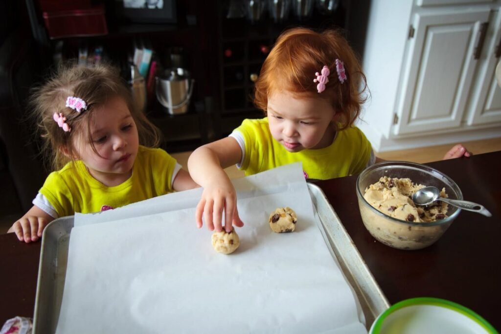 Two children in yellow shirts carefully placing dough balls on a parchment-lined tray. A bowl of cookie mix with a spoon sits nearby in the kitchen.