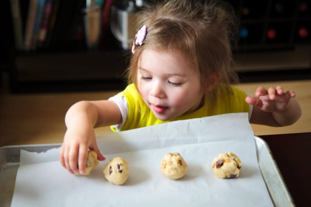 A young child in a yellow shirt carefully places cookie dough balls on a parchment-lined tray.