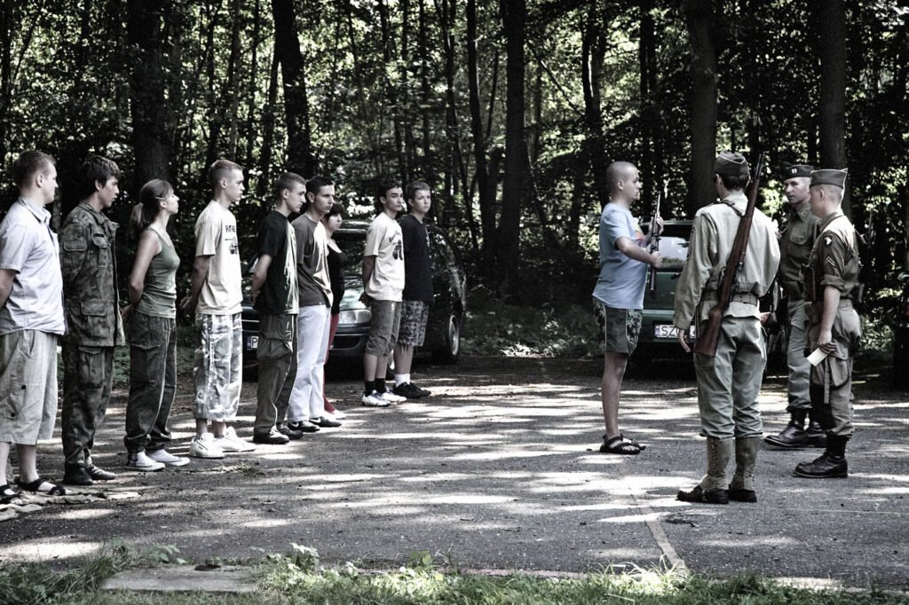 A group of military personnel and civilians gathered in a forest clearing for a meeting or brief.
