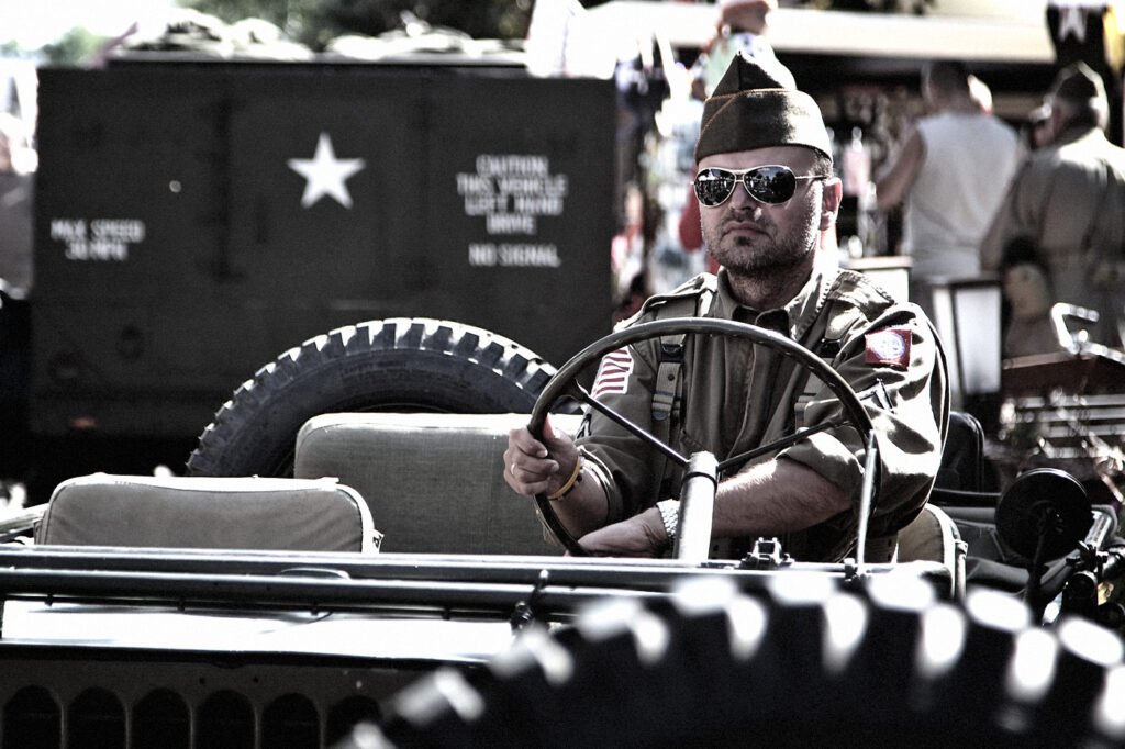 A man in military uniform sits behind the wheel of an antique military vehicle at an outdoor event.