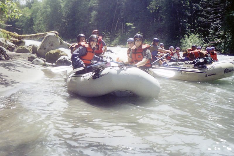 A group of people in orange life jackets and helmets are enjoying a rafting trip on a river surrounded by lush green forest.