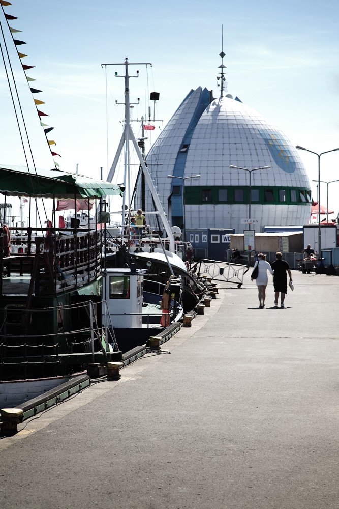 A couple strolling on a sunny day along the paved walkway of the London Docklands pier. The iconic dome of the Museum of London Docklands is prominently visible in the background.