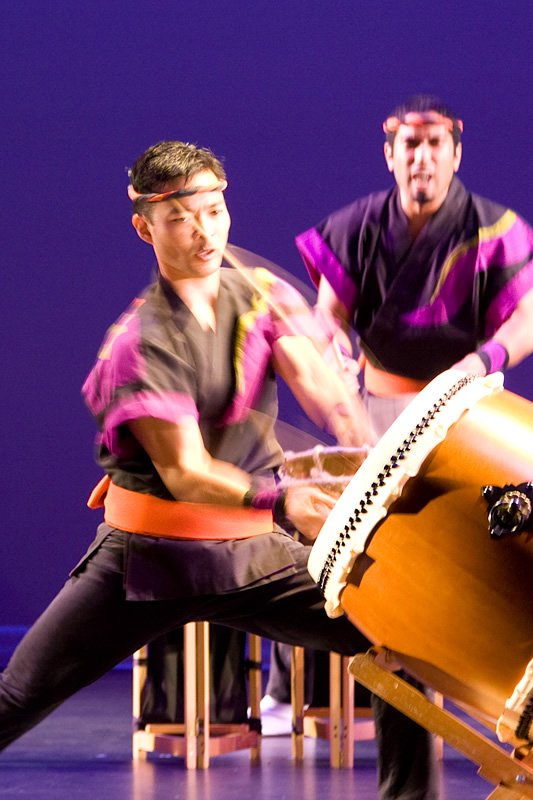 Two performers dressed in vibrant costumes energetically play traditional taiko drums on stage.