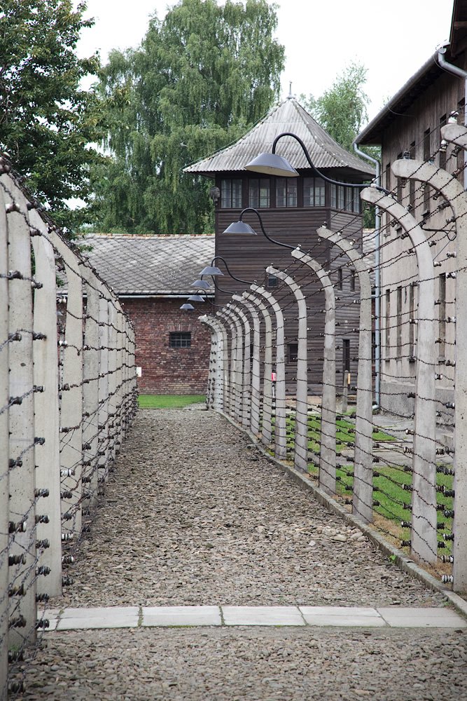 A path bordered by barbed wire fences leading between wooden barracks and a watchtower at the Auschwitz concentration camp.
