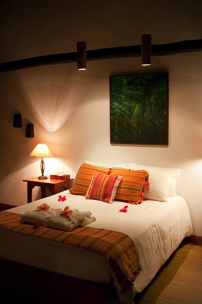 A cozy bedroom features warm lighting, a neatly made bed with orange and white pillows, and a small bedside table with a lamp. A piece of art hangs above the bed, adding a touch of elegance to the room.