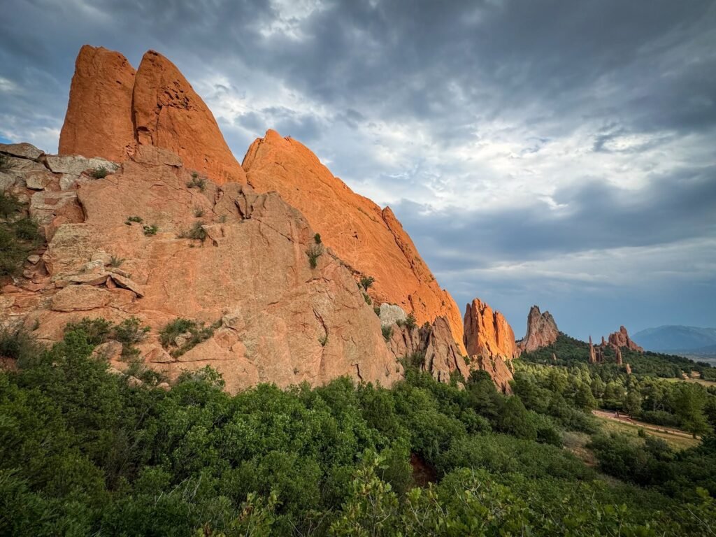 Striking red sandstone spires rise against a cloudy blue sky above lush green vegetation in Garden of the Gods, near Colorado Springs.