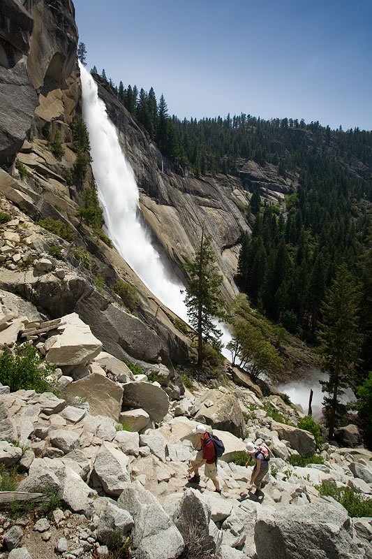 Two hikers are trekking through rocky terrain near a stunning waterfall. The waterfall cascades down from a high cliff, surrounded by lush greenery.