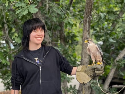 Kristie smiles while holding a falcon on her gloved hand, surrounded by green trees in a natural outdoor setting.