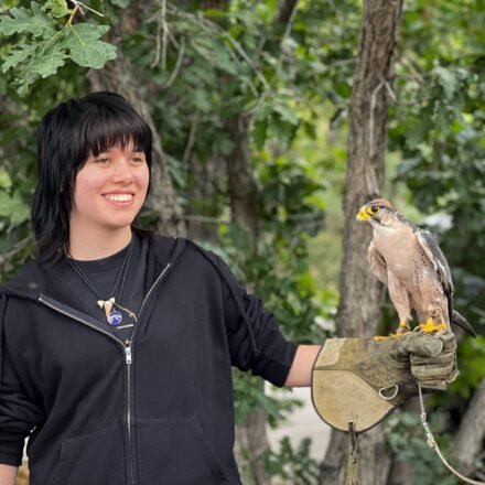 Kristie smiles while holding a falcon on her gloved hand, surrounded by green trees in a natural outdoor setting.