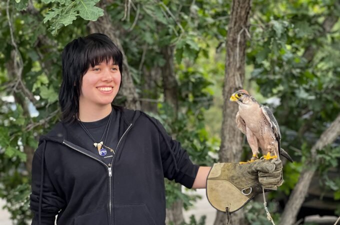 Kristie smiles while holding a falcon on her gloved hand, surrounded by green trees in a natural outdoor setting.