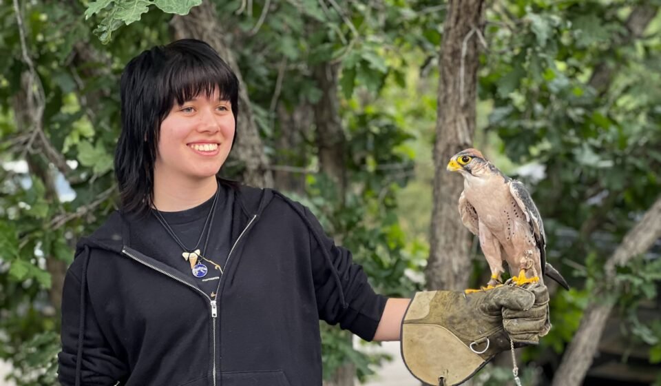 Kristie smiles while holding a falcon on her gloved hand, surrounded by green trees in a natural outdoor setting.