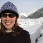 Kanye West Kristie is smiling brightly, wearing a blue patterned beanie and sunglasses, standing in a snowy landscape with mountains in the background.