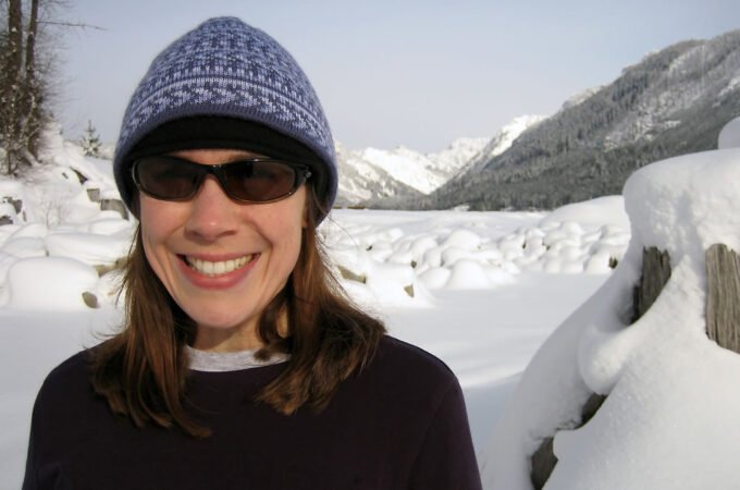 Kristie is smiling brightly, wearing a blue patterned beanie and sunglasses, standing in a snowy landscape with mountains in the background.
