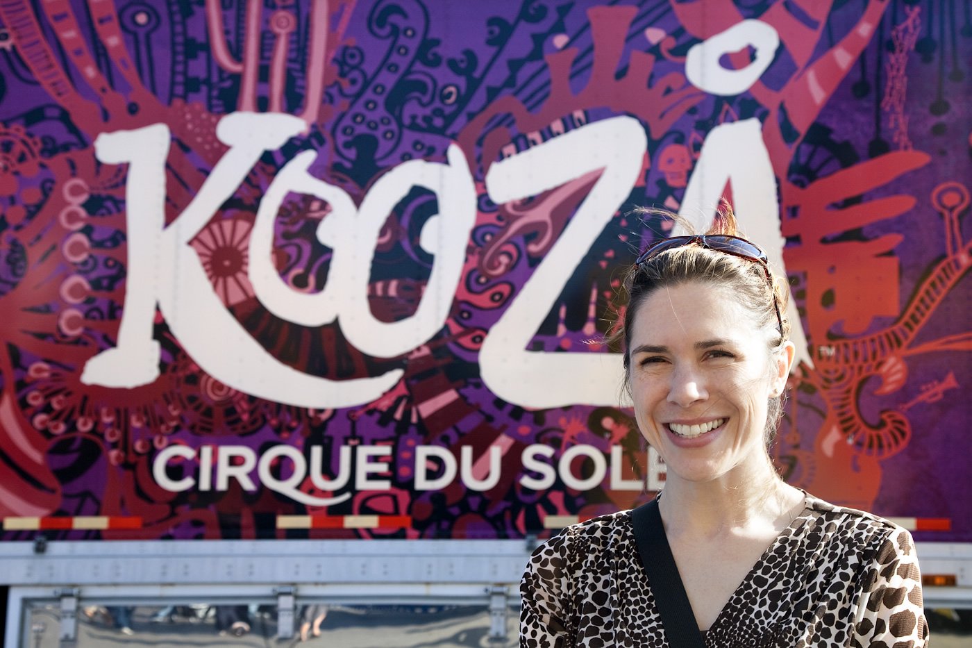 A woman stands smiling in front of a vibrant KooZA Rawhauh by Cirque du Soleil sign.