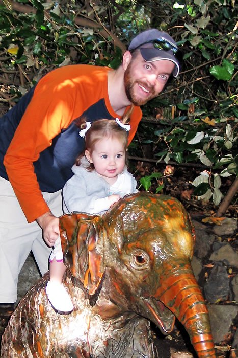 John holding a toddler while exploring outdoors near an elephant statue, surrounded by lush greenery.