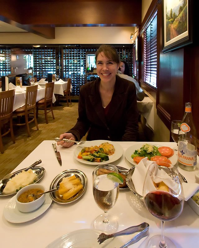 A woman smiles at a dining table filled with various dishes and drinks in a cozy restaurant setting.