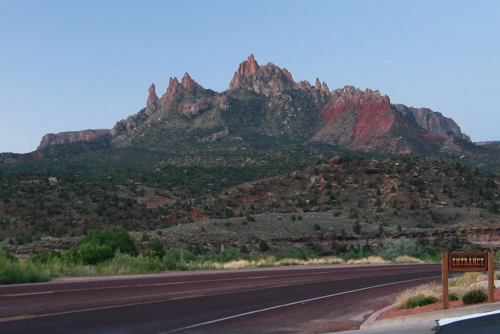 A beautiful landscape featuring towering red rock formations under a clear blue sky. A road runs along the bottom with a signpost on the right side.